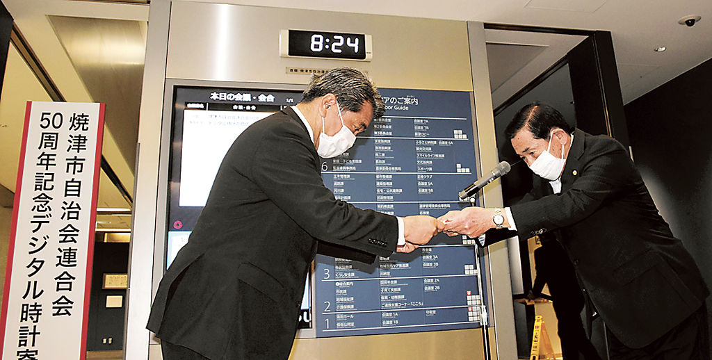 デジタル時計の下で目録を手渡す岩崎会長（右）＝焼津市役所