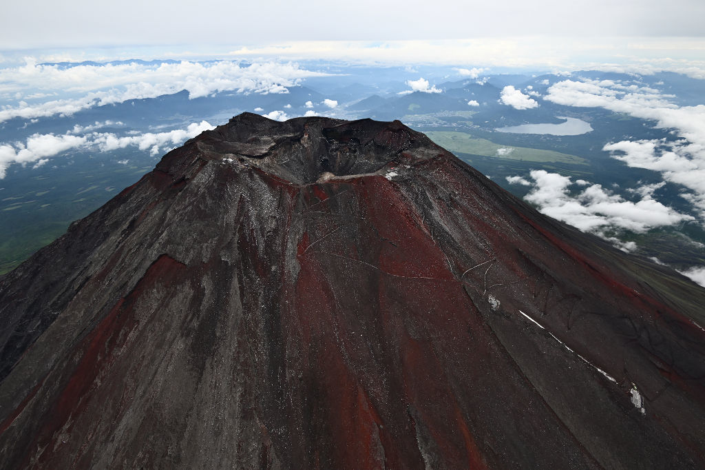 赤い山肌を見せる夏の富士山。山頂付近はうっすらと雪に覆われた。右下は臨時支局を開設した万年雪山荘＝１日午前（静岡新聞社ヘリ「ジェリコ１号」から、写真部・久保田竜平）