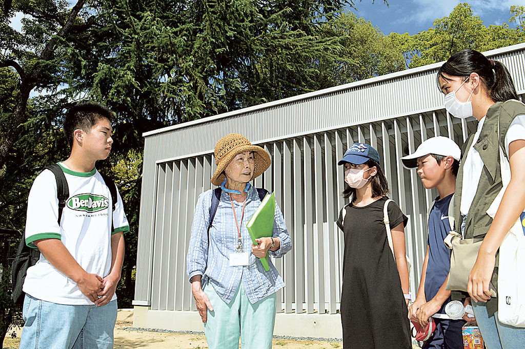 金本隆子さん（左から２人目）の説明に聞き入る山本惇太さん（左）ら森町の児童生徒＝５日午後、広島市中区の平和記念公園